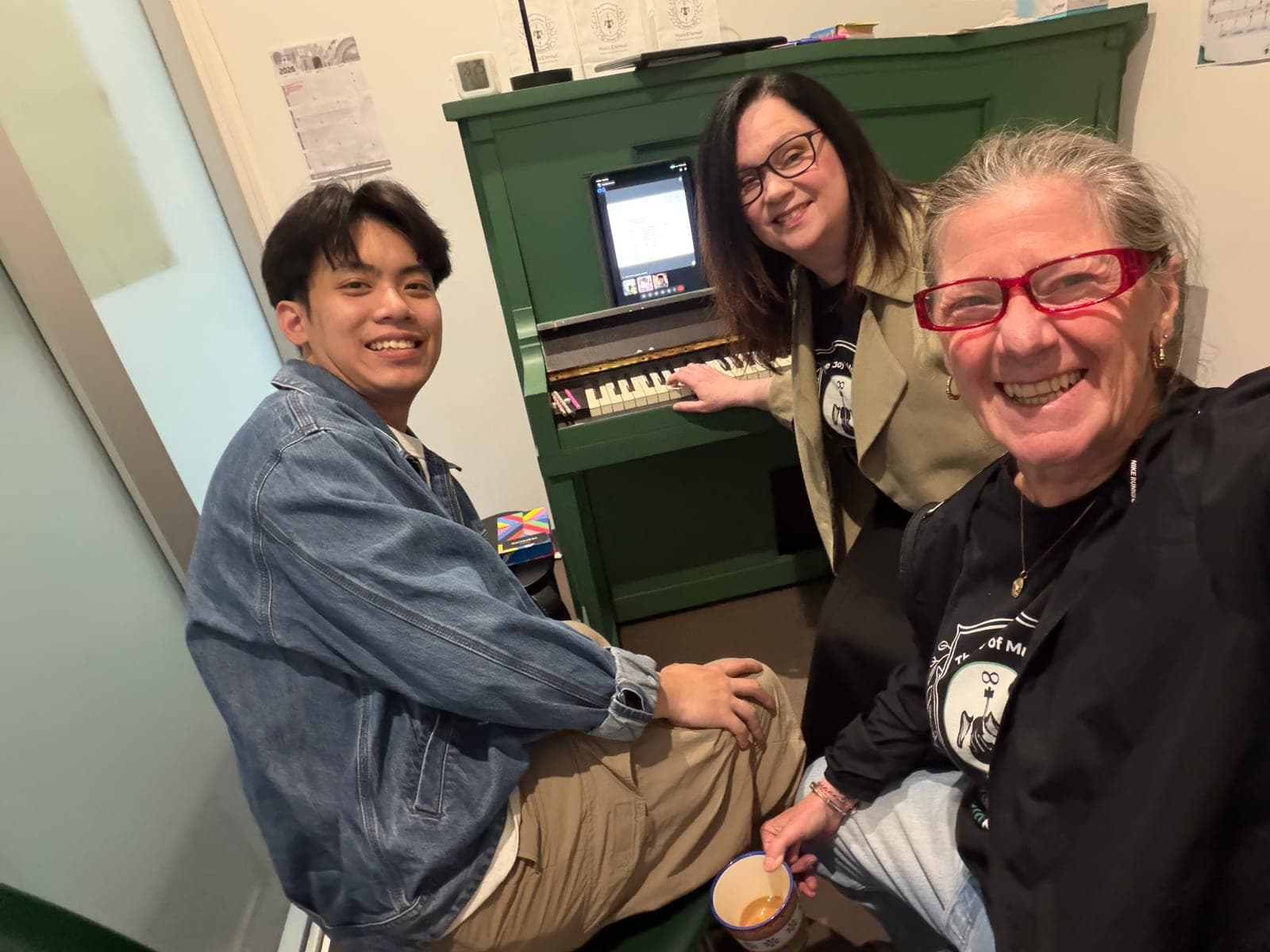 Three people gathered around the green upright piano in the Music Eternal studio, one playing, coffee in hand