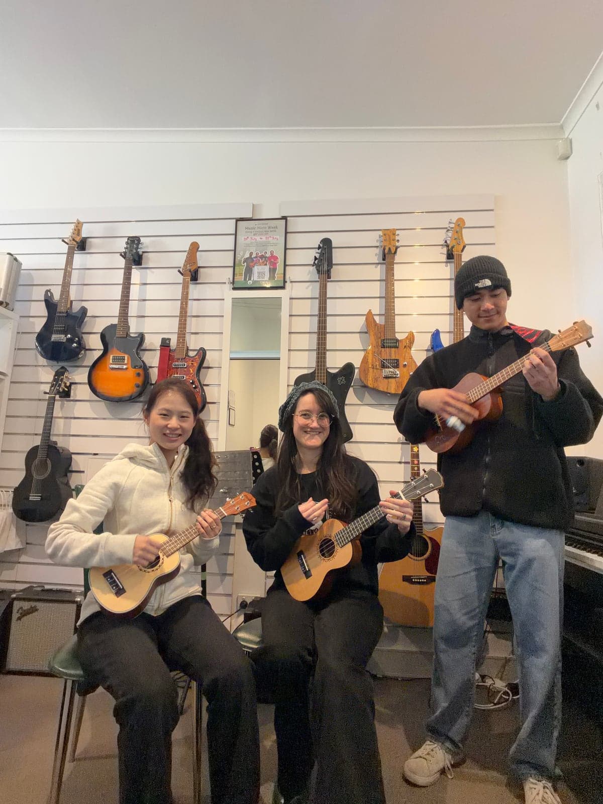 Three people holding ukuleles in front of the guitar wall at the Music Eternal studio